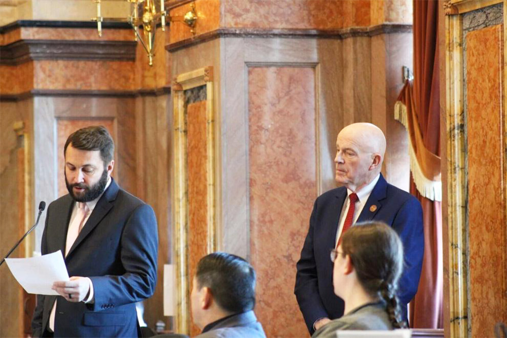 Two people in suits stand at a podium inside an ornate chamber, with one reading from a paper while others sit nearby.