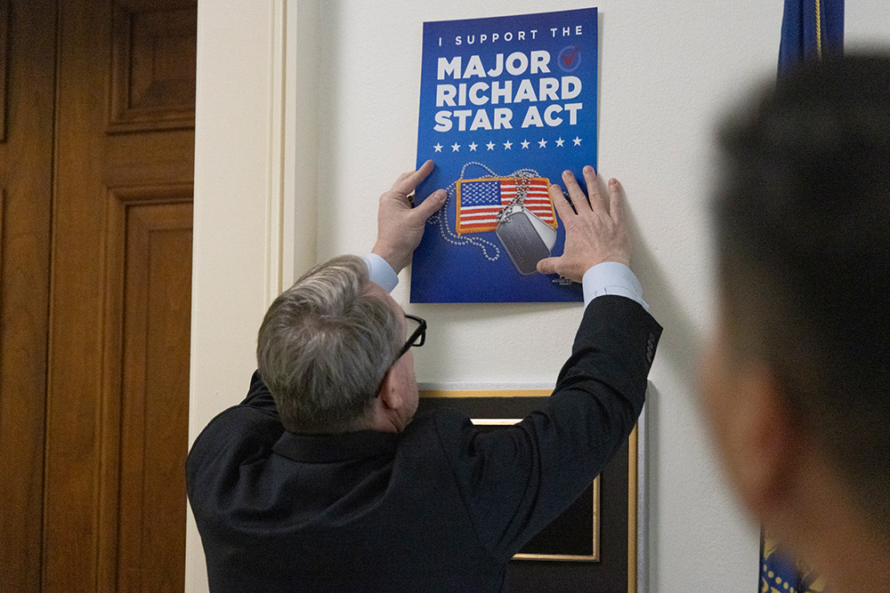 A WWP staff member hangs a poster on a wall that says “I support the Major Richard Star Act,” and includes a picture of an American flag and dogtags. 