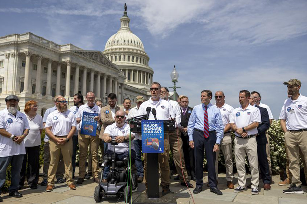 Group of advocates stand at a podium outside the U.S. Capitol during a press event, holding signs supporting the Major Richard Star Act.
