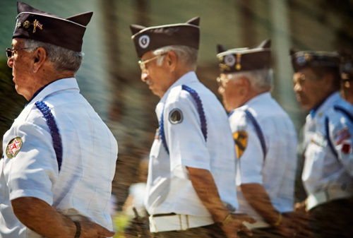Uniformed veterans in white shirts and caps stand in a line outdoors during a ceremonial event, viewed from the side.