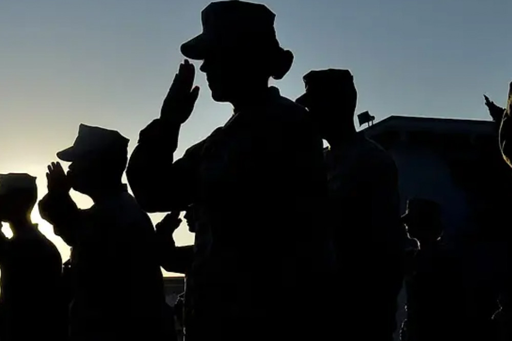 Silhouetted uniformed service members stand in formation, raising their hands in a salute against a bright sky.