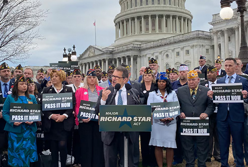 Large group gathered outside the U.S. Capitol at a press rally, with speakers at a podium and attendees holding signs reading “Major Richard Star Act — Pass It Now.”