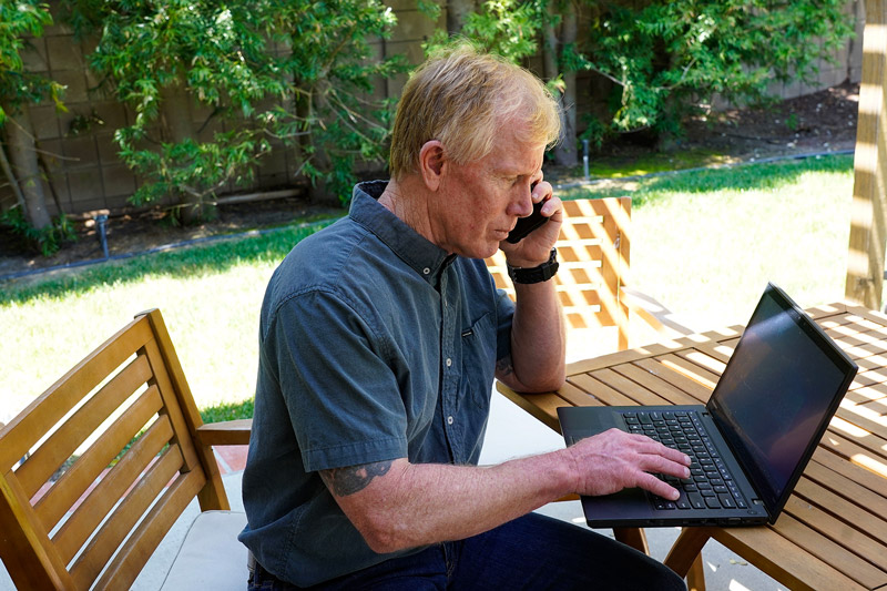 Man sitting on a patio at a wooden table using a smartphone and laptop.