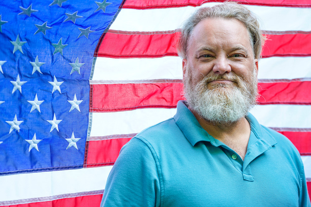 A veteran wearing a green polo shirt is smiling while standing in front of an American flag.