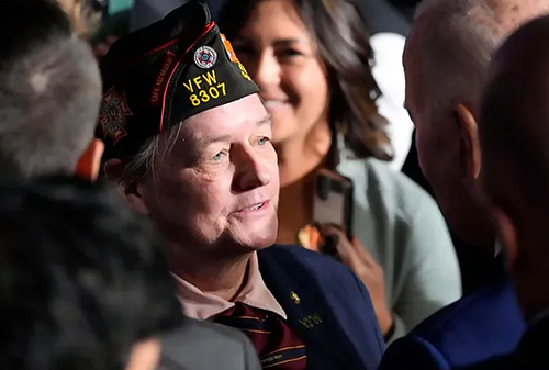 Close-up of a veteran wearing a VFW cap speaking with others in a crowded indoor setting, surrounded by people and cameras.