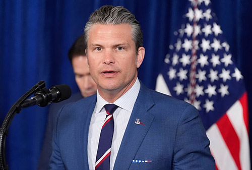 Defense Secretary Pete Hegseth speaking at a podium with microphones, wearing a blue suit and striped tie, with a U.S. flag visible in the background.