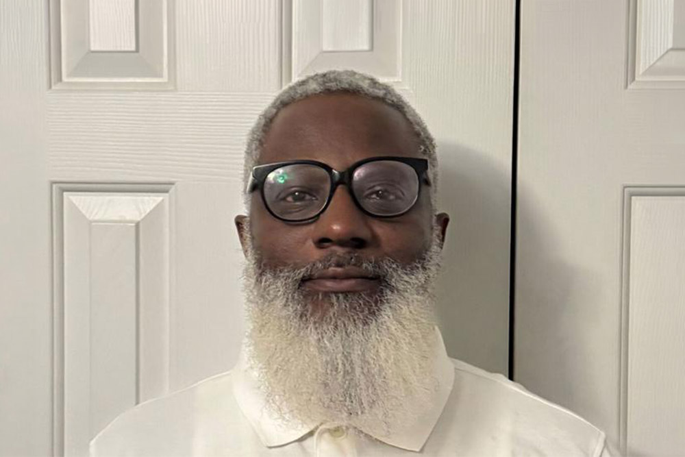 Head-and-shoulders portrait of a person wearing round glasses and a white shirt, standing in front of a white paneled door.