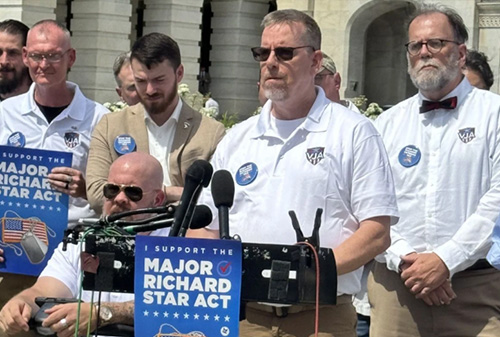 Close-up of speakers and supporters standing behind microphones at an outdoor press event, with signs reading “Support the Major Richard Star Act.”