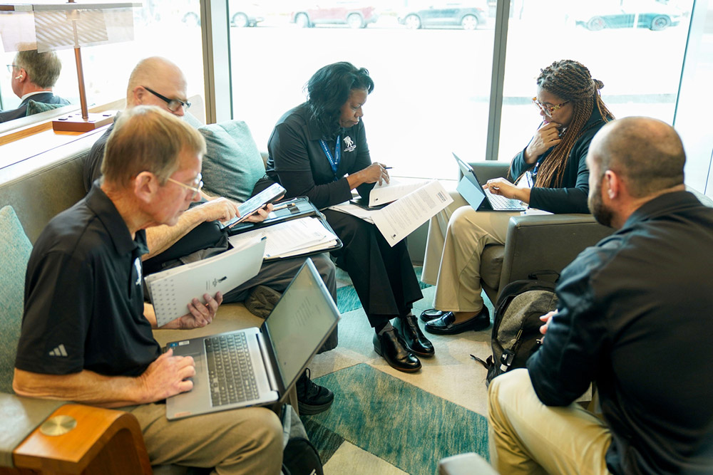 A small group of people sit in a lounge area collaborating with laptops and printed documents near a large window.