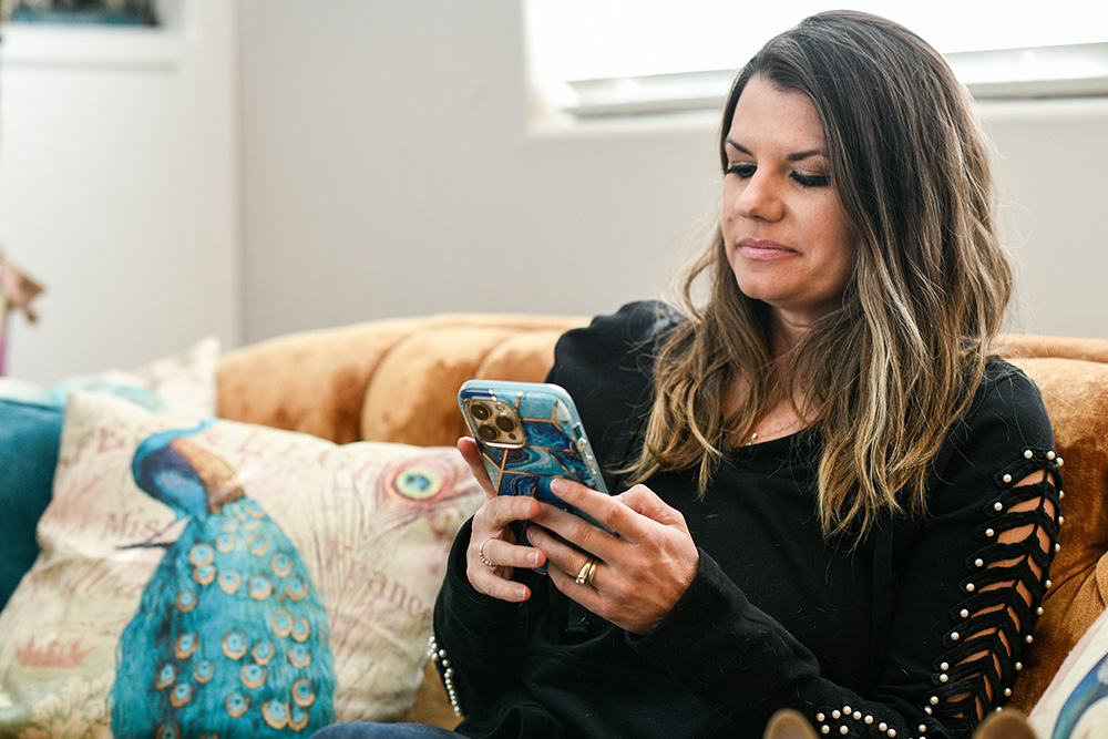 Veteran Melissa McMahon sits on her sofa while holding her cell phone in both hands and looking at the screen.